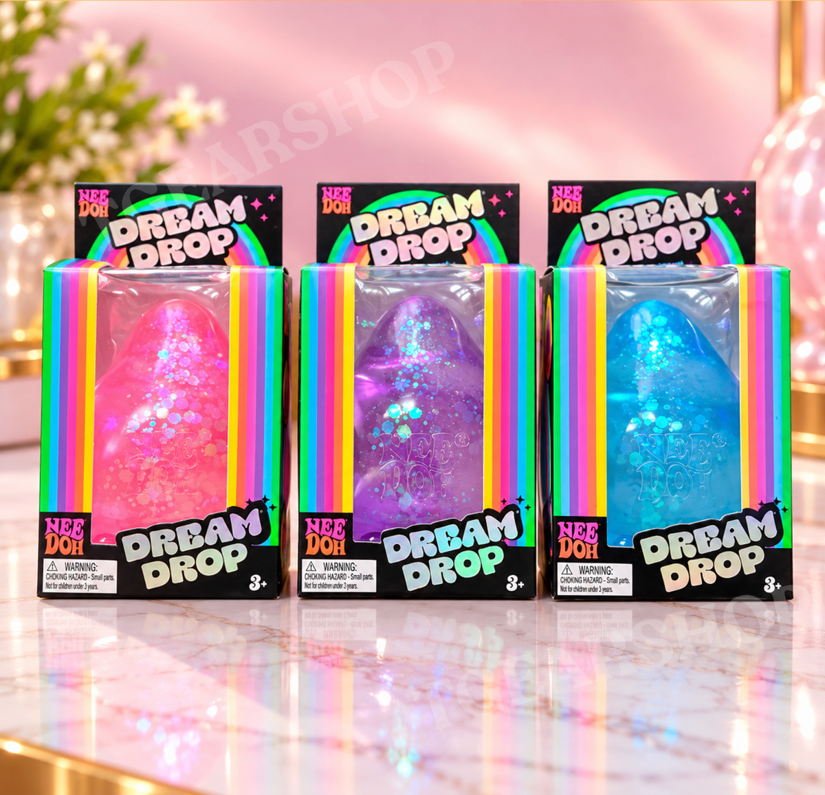 Three NeeDoh Dream Drop squishies in pink, purple and blue, each in its own rainbow-trimmed retail packaging.